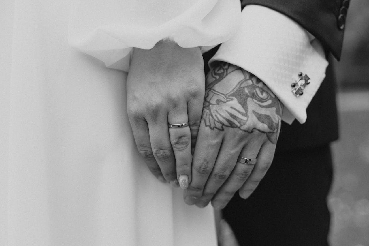 heros-img Close-up shot of married couple holding hands with rings, showcasing tattoos.